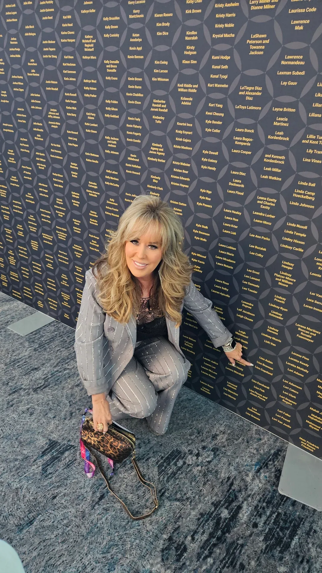 Lisa Copeland in a pinstriped blazer kneeling and pointing at a donor wall filled with names