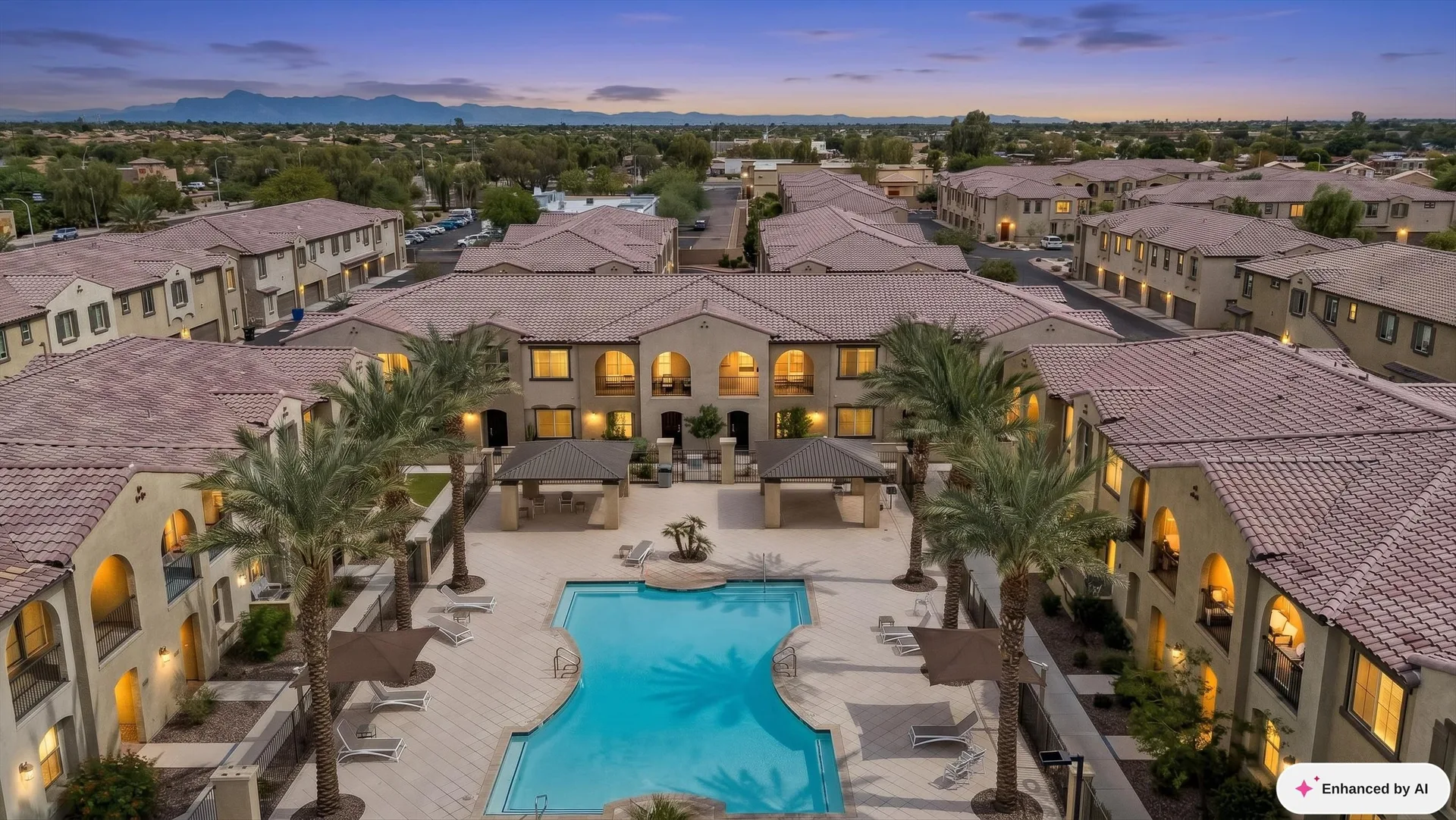 Twilight aerial view of a multi-family apartment community with a central swimming pool, palm trees, and residential buildings