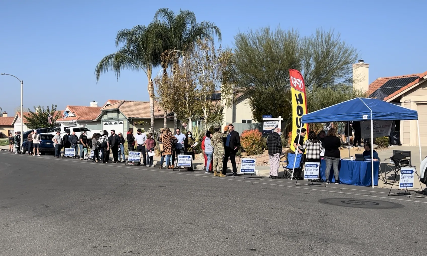 Homebuyers lined up outside an open house under a blue tent with signage