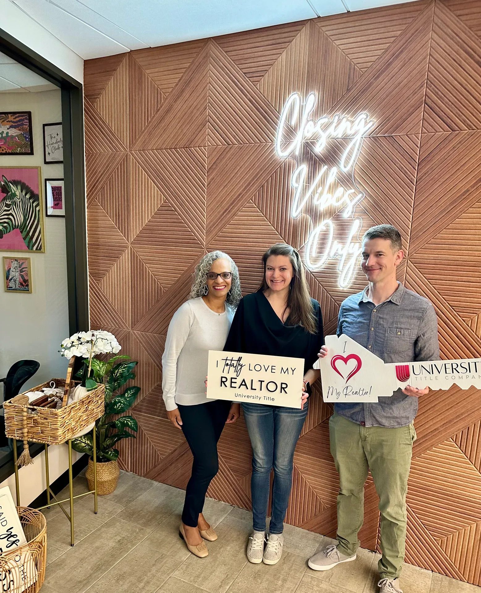 Tami Price with two clients holding closing props in front of a wooden wall with a neon sign