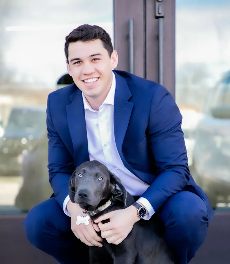 Justin Murillo in a navy blazer, crouching and smiling while holding his silver Lab puppy, Murphy