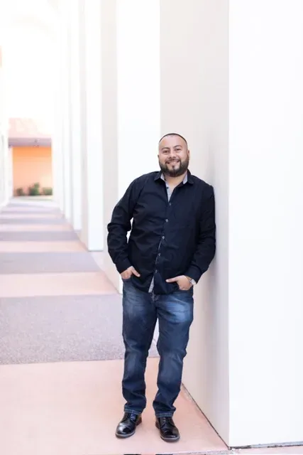 Ernesto Ramirez in a black button-down shirt and dark jeans, leaning against a white pillar