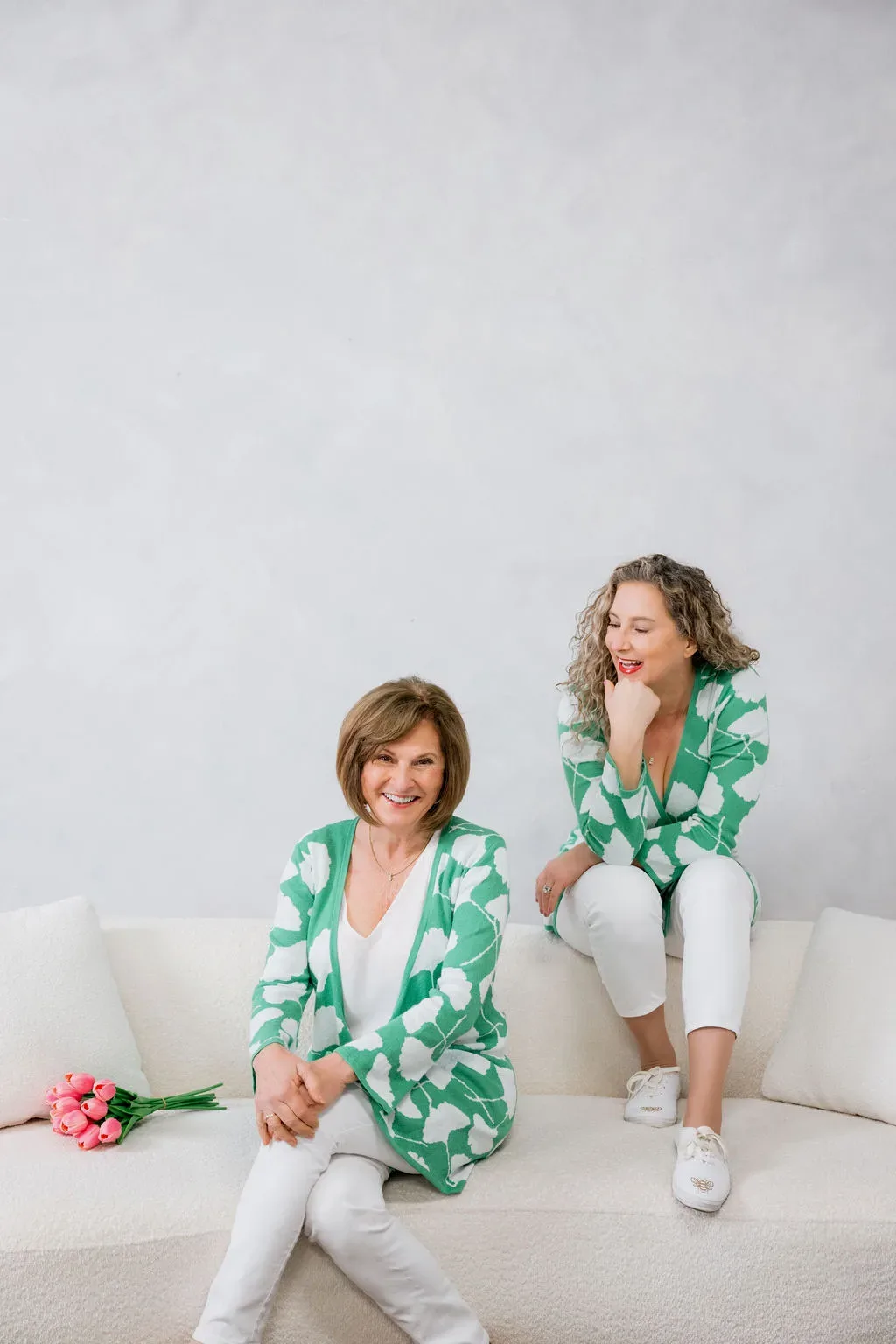 Two women in matching green cardigans sitting on a white sofa with pink flowers nearby
