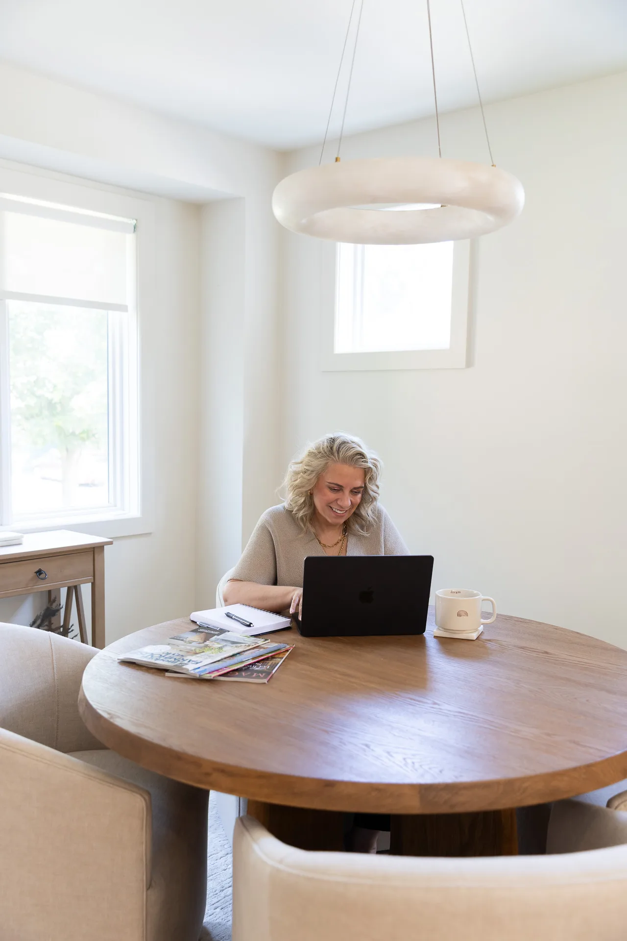 Kelly MacDonald-Wilson smiling while working on her laptop at a round wooden table in a bright, modern home office
