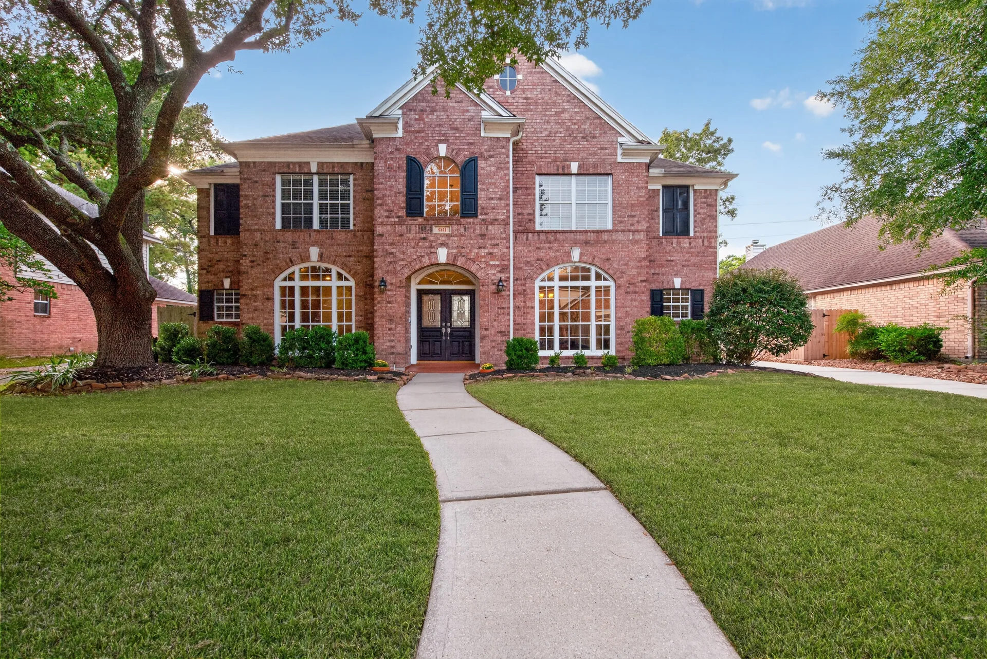 Front exterior of 4211 Brook Shadow Dr, a two-story brick home with arched windows, black shutters, and a mature tree in the yard