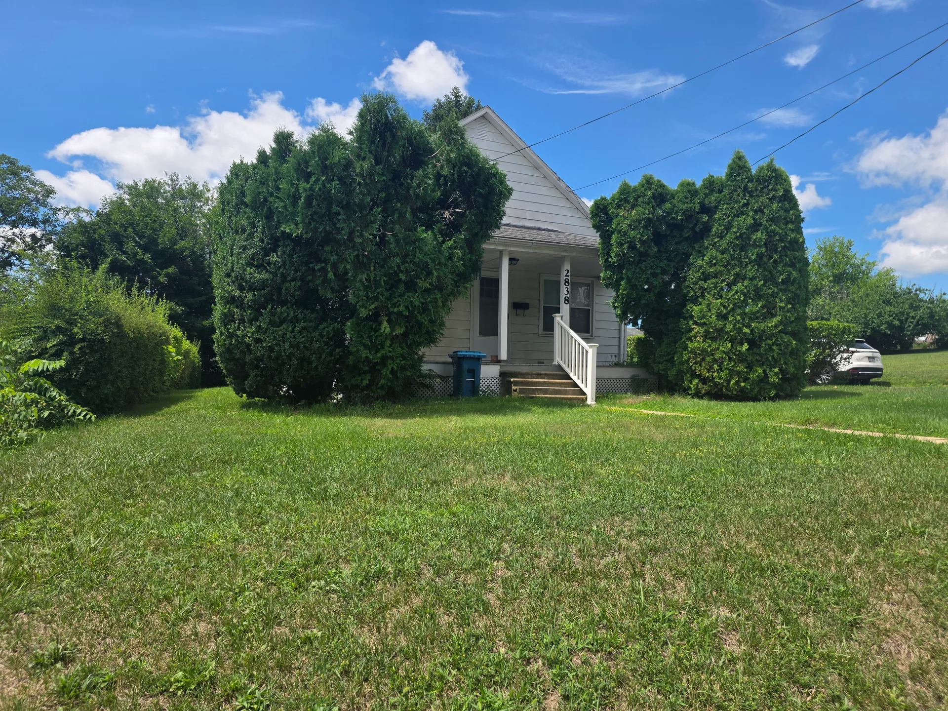 Street-level view with covered front porch, white railings, and evergreen trees